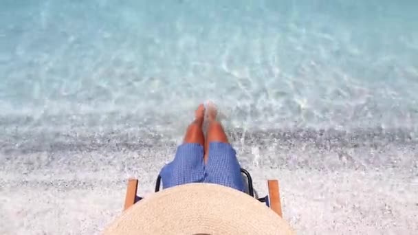 Fiatal nő a kalapban ül a tengerparti széken, és rázza a lábát a fantasztikus kék tengerbe. Nyaralási koncepció. — Stock videók