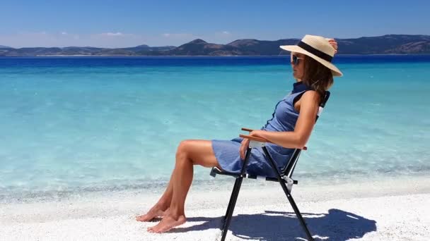 Mujer joven en el sombrero se sienta en la silla de playa y se broncea cerca de los océanos cristalinos de agua y el mar azul. Concepto de vacaciones. — Vídeos de Stock