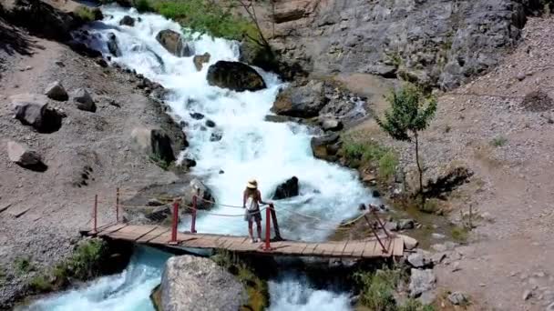AERIAL. Kvinnlig turist står på bron nära enorma vattenfall. Kvinna tittar till källorna bergen floden, utsikt från drönaren. — Stockvideo