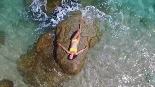 바다에 있는 바위에 누워 있는 노란색 비키니를 입은 젊고 아름다운 패션 모델. 패션 촬영. 태국에서의 항공 영상. 휴가를 즐기며 — 비디오
