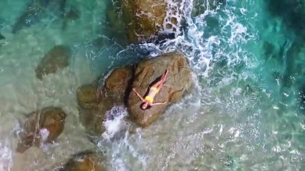 Joven y hermosa modelo femenina de moda en bikini amarillo que yace sobre la roca en el mar. Disparo de moda. Video aéreo en la isla de Tailandia. Relajarse de vacaciones — Vídeos de Stock