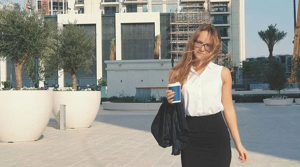 Jeune femme d'affaires souriante dans des lunettes et une tasse de café marchant dans la ville avec des bâtiments modernes sur le fond. — Photo