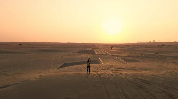 Πίσω όψη ξανθά μαλλιά γυναίκα με κίτρινο αυθεντικό σακίδιο ψάχνει σε αμμώδη δρόμο μετά την αμμοθύελλα. Ταξίδια περιπέτειες στην έρημο. — Φωτογραφία Αρχείου