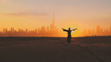 Geleneksel BAE elbisesi giymiş Arap bir kadın. Arkasında Dubai şehir silueti olan bir çölde gün batımında Abean ellerini kaldırıyor..