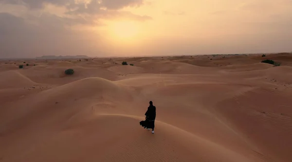 AERIAL. Câmera seguindo mulher no vestido tradicional Emirati andando em um deserto no vento strog e pôr do sol. — Fotografia de Stock