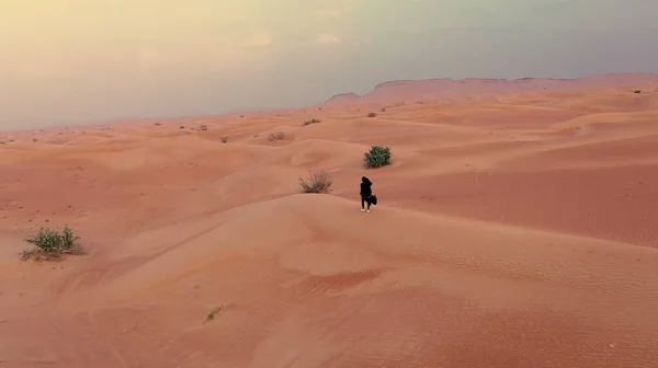 AERIAL. Câmera seguindo mulher no vestido tradicional Emirati andando em um deserto no vento strog e pôr do sol. — Fotografia de Stock