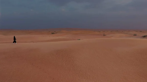 AERIAL.身着传统阿联酋服装的女人在沙漠中迎风而行，夕阳西下，相机紧跟在她后面. — 图库照片