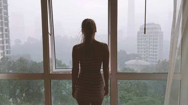 Vista posteriore della donna che guarda attraverso la finestra sulla città sotto la pioggia. — Foto Stock