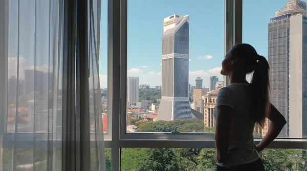 Женщина открывает занавес на больших окнах с видом на город с зеленым гадерном и зданиями. Концепция зеленого города. — стоковое фото