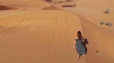 AERİAL. Dubai çölünde gün batımında kumulda yürüyen uzun elbiseli bir kadın.