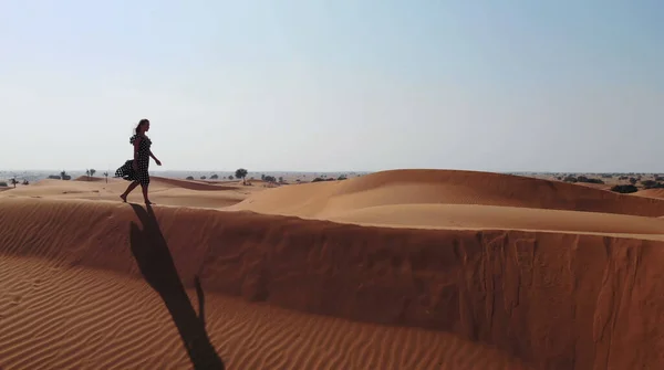 AERIAL. Женщина в длинном платье, прогуливаясь по песчаным дюнам пустыни Дубая со следами на песке во время заката — стоковое фото