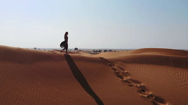 AERIAL. Женщина в длинном платье, прогуливаясь по песчаным дюнам пустыни Дубая со следами на песке во время заката — стоковое фото