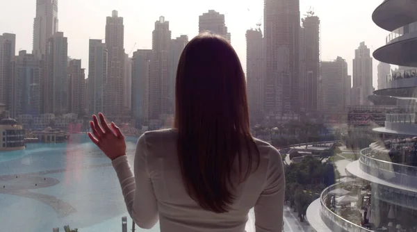 Mujer joven tomando fotos en la ventana grande y mirando a los edificios de la ciudad de Dubai. Puesta de sol. — Foto de Stock