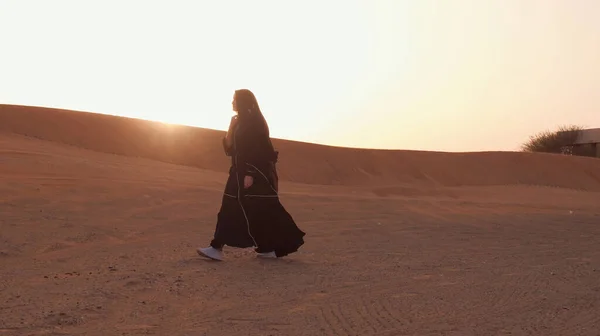 Retrato de uma jovem árabe vestindo roupas pretas tradicionais durante o belo pôr do sol sobre o deserto. — Fotografia de Stock