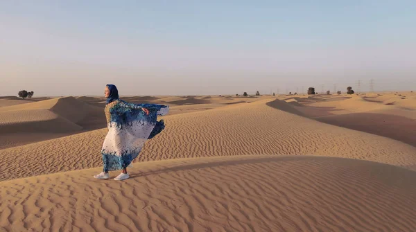 Повітряний вид з дрона, який летить поруч з жінкою в традиційному одязі Абая Об'єднаних Арабських Еміратів, що йде по дюнах в пустелі Пустельний квартал. Абу-Дабі, ОАЕ. — стокове фото