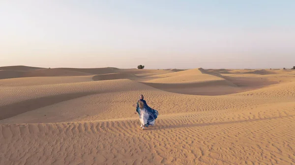 Повітряний вид з дрона, який летить поруч з жінкою в традиційному одязі Абая Об'єднаних Арабських Еміратів, що йде по дюнах в пустелі Пустельний квартал. Абу-Дабі, ОАЕ. — стокове фото