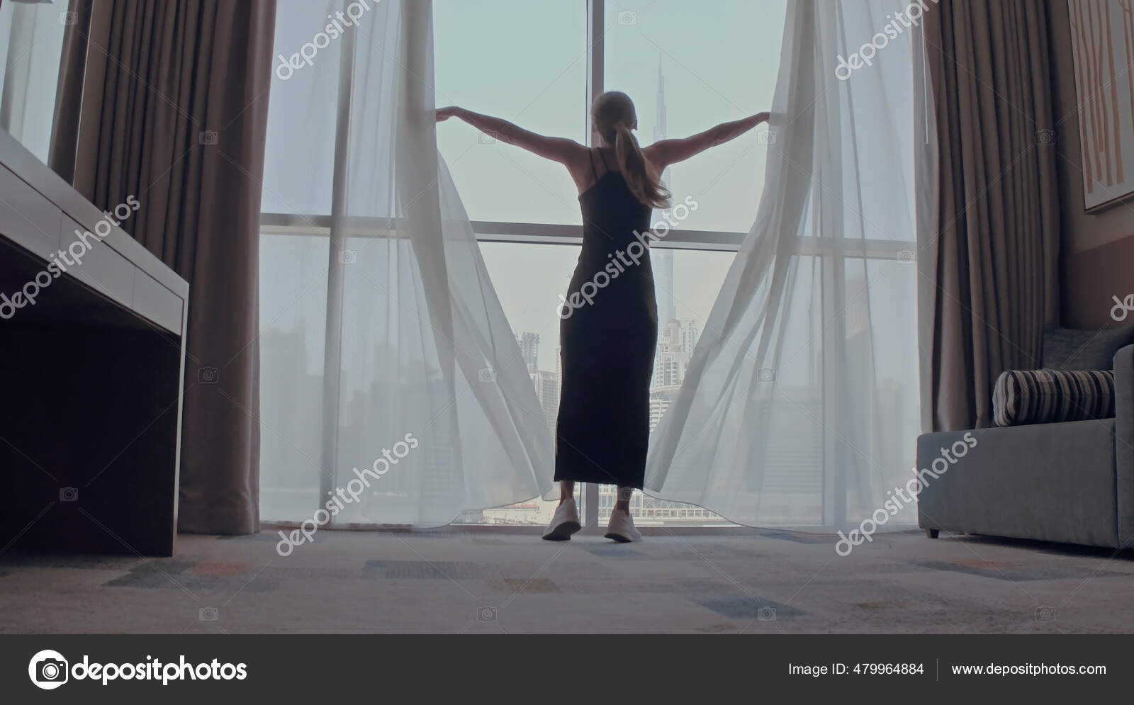 Back view of woman opening curtains and looking out of window to