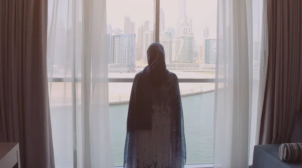 Vista trasera de la mujer abriendo cortinas y mirando por la ventana al paisaje urbano en el hotel por la mañana. Edificios modernos en la ciudad de Dubai — Foto de Stock