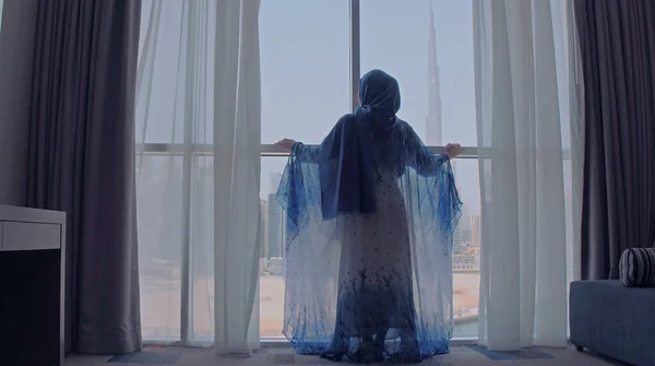 Visão traseira da mulher abrindo cortinas e olhando de janela em paisagem urbana no hotel pela manhã. Edifícios modernos na cidade de Dubai — Fotografia de Stock