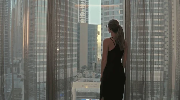 Silueta de mujer joven abre cortinas en la gran ventana extiende los brazos y mirando a su apartamento en los edificios de la ciudad durante el increíble amanecer. Dubai ciudad — Foto de Stock