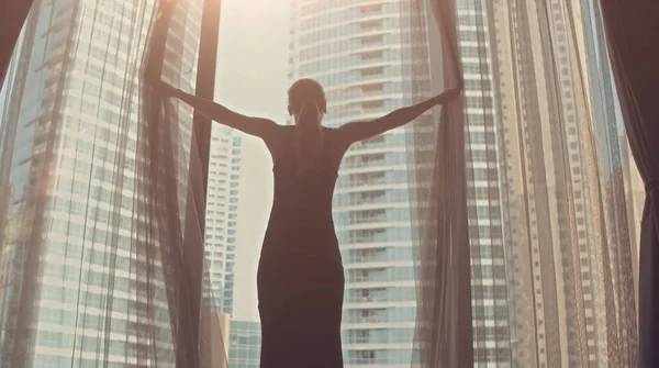 Genç kadın silueti büyük pencerenin perdelerini açıyor kollarını açıyor ve gün doğumunda apartmandaki apartmanından dışarı bakıyor. Dubai şehri