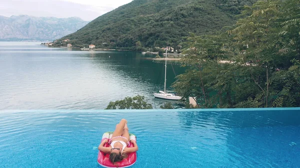 AÉRIAL. Charmante jeune femme caucasienne blonde en lunettes de soleil et maillot de bain nage sur un matelas gonflable dans la piscine avec vue magnifique sur la mer — Photo