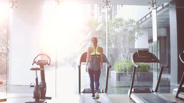 Koşu bandında yürüyen bir kadın panoramik bir pencerede ve sabahın köründe spor salonunda güneş doğuyor..