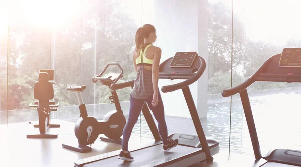 Koşu bandında yürüyen bir kadın panoramik bir pencerede ve sabahın köründe spor salonunda güneş doğuyor..