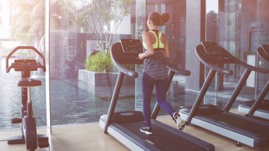 Koşu bandında yürüyen bir kadın panoramik bir pencerede ve sabahın köründe spor salonunda güneş doğuyor..