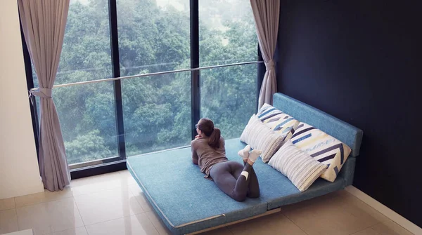 Asyalı iş kadını gündelik kıyafetlerini giyiyor. Oturma odasında kanepede dinleniyor. Yaz tatili seyahati, pencerenin önünde yağmur..