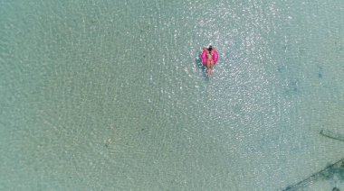 Şeffaf mavi denizde şişirilebilir büyük donutta yüzen şapkalı güzel bir kadının üst görüntüsü. Yüzen yatakta dinlenen bayanın hava görüntüsü..