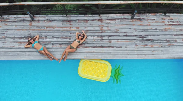两个女人躺在游泳池边，有一个松果形状的充气床垫，俯瞰天空。在海滩玩乐和放松的假期. — 图库照片