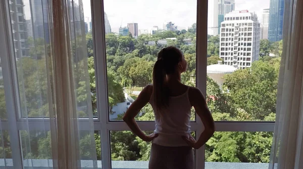 Mujer joven destapar la gran ventana y mirando por su apartamento en los edificios de la ciudad — Foto de Stock