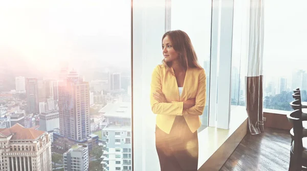 Sarı ceketli genç bir kadın şehrin arka planında pencerenin önünde duruyor..