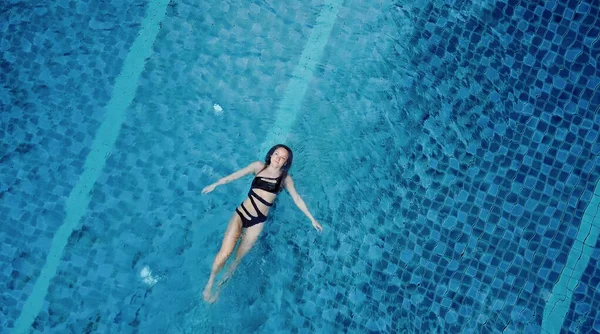 Верхний вид воздушной фотографии соблазнительной сексуальной модели в черном купальнике наслаждается отдыхом в бассейне отеля. Потрясающая модная женщина в бикини, отдыхающая в роскошном спа — стоковое фото
