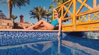 fit vücutlu, göbekli, masa başında havuz kenarında uzanan ve güneşlenen güzel beyaz bikinili kadın. Kadın havuz başında dinleniyor ve güneşli yaz gününün tadını çıkarıyor.