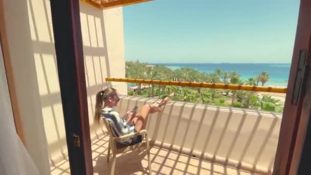 Widok z tyłu kobiety siedzi na krześle na balkonie z widokiem na morze i za pomocą smartfona. — Wideo stockowe