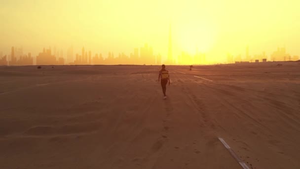 AERIAL. Молодая женщина-путешественница стоит на дороге после песчаной бури, глядя на силуэт города Дубая и фотографируется на смартфоне. — стоковое видео