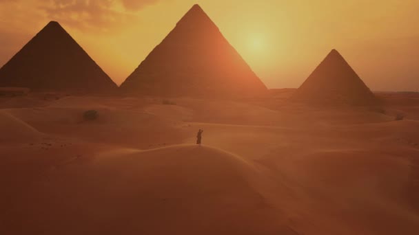 Au ralenti. Silhouette de femme en robe arabe traditionnelle lève les bras au coucher du soleil avec des pyramides sur le fond. — Video