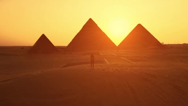 Mujer viajera mira a la silueta de las pirámides al atardecer en el desierto. Disparo aéreo. — Vídeos de Stock