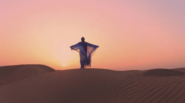 Mulher árabe vestida em vestido tradicional dos Emirados Árabes Unidos abayain levantando as mãos no pôr do sol em um deserto. — Fotografia de Stock