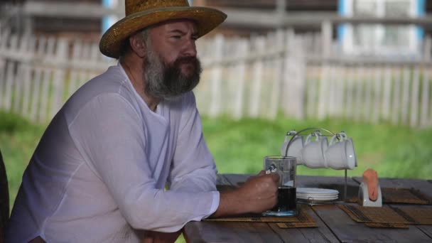 Farmer, uomo con la barba in un cappello di paglia, si siede a un tavolo e beve birra scura da una tazza di vetro — Video Stock