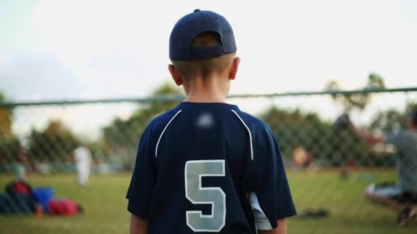 Niño pequeño en uniforme de béisbol observando la práctica detrás de la valla en el campo — Vídeo de stock