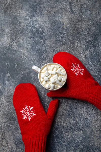 Natal e Ano Novo composição de férias acolhedora com cachecol, mãos de mulher em mitenes, canecas com bebida quente e marshmallow no fundo de concreto cinza. Deitado plano, vista superior. — Fotografia de Stock