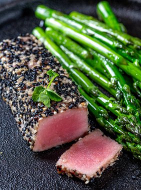 Siyah susamlı kızarmış ton balığı bifteği ve siyah tabakta kuşkonmaz.