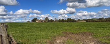 Uzun bir açık çayır tarlası mavi gökyüzü ufkuna ve yavaş hareket eden bulutlara doğru uzanıyor. Uzak köşede, Horn Lane, Silsden, Yorkshire, İngiltere yakınlarındaki dağınık ağaçların arasında yalnız bir çiftlik evi yuva yapar.