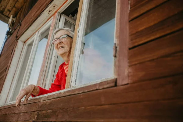 사랑스럽고, 미소짓고 있는 할머니가 창밖을 내다보며 화창 한 날, 은퇴 후의 삶을 즐기고 있습니다 — 스톡 사진