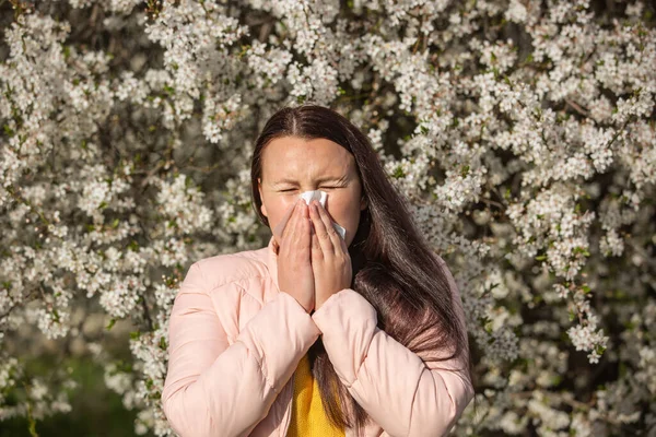 Concepto de síntomas de alergia de primavera, mujer joven estornudando frente a la floración de un árbol, alergia al polen y la temporada de floración, atención médica — Foto de Stock