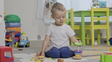 Yerde oturan sevimli küçük çocuk oyun odasında renkli ahşap bir piramit oyuncağı yapıyor.
