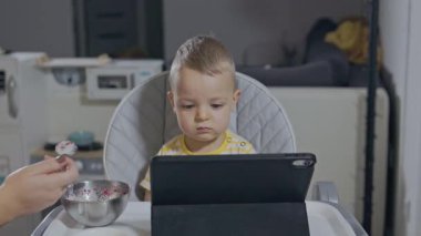 Genç anne oğlunu yüksek sandalyede beslerken o dijital tablette çizgi film izliyor.
